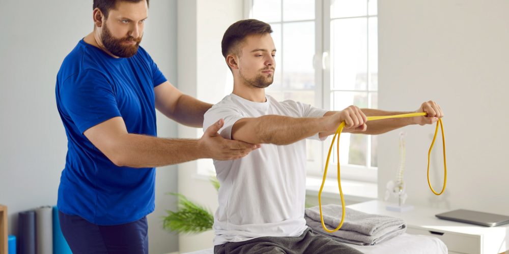 Physiotherapy specialist and his male patient use special equipment during rehabilitation. Young man doing remedial physical exercises with resistance band under supervision of serious physiotherapist